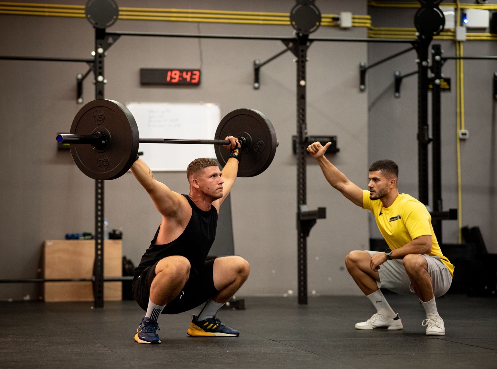 Athlete performing weightlifting exercise under trainer's guidance in a gym setting.